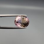 ​A precision-faceted 5.36-carat oval natural Ametrine gemstone held in tweezers against a gray backdrop, displaying a sharp half-and-half color partition of purple amethyst and golden-amber citrine with brilliant clarity.