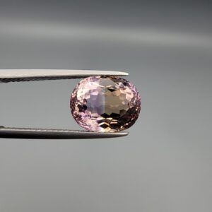 ​A precision-faceted 5.36-carat oval natural Ametrine gemstone held in tweezers against a gray backdrop, displaying a sharp half-and-half color partition of purple amethyst and golden-amber citrine with brilliant clarity.