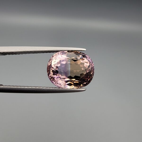 ​A precision-faceted 5.36-carat oval natural Ametrine gemstone held in tweezers against a gray backdrop, displaying a sharp half-and-half color partition of purple amethyst and golden-amber citrine with brilliant clarity.