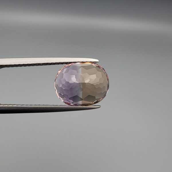 ​A precision-faceted 5.36-carat oval natural Ametrine gemstone held in tweezers against a gray backdrop, displaying a sharp half-and-half color partition of purple amethyst and golden-amber citrine with brilliant clarity.