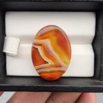 A high-gloss 31.42-carat oval natural Yemeni carnelian banded agate cabochon resting in a black jewelry box. The stone displays rich carnelian red, sharp white fortification banding, honey yellow, and dark brown-black layers with impressive clarity and polish.