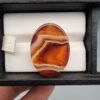 A high-gloss 31.42-carat oval natural Yemeni carnelian banded agate cabochon resting in a black jewelry box. The stone displays rich carnelian red, sharp white fortification banding, honey yellow, and dark brown-black layers with impressive clarity and polish.