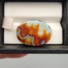 A 27.57-carat oval natural Yemeni carnelian agate cabochon shown in a black display case. The front side displays vibrant botryoidal orange, red, yellow, and white cloud patterns, while the back shows a dark blackish base with an illuminated fiery orange and red internal cloud core.