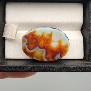 A 27.57-carat oval natural Yemeni carnelian agate cabochon shown in a black display case. The front side displays vibrant botryoidal orange, red, yellow, and white cloud patterns, while the back shows a dark blackish base with an illuminated fiery orange and red internal cloud core.