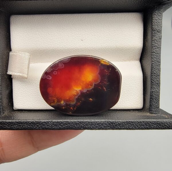 A 27.57-carat oval natural Yemeni carnelian agate cabochon shown in a black display case. The front side displays vibrant botryoidal orange, red, yellow, and white cloud patterns, while the back shows a dark blackish base with an illuminated fiery orange and red internal cloud core.