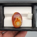 "Natural Carnelian Banded Agate Sulemani Aqeeq gemstone 32.29 carat oval cabochon from Yemen held in tweezers showing red, orange, yellow, and white natural color bands"