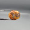 A 10.79-carat oval natural Yemeni carnelian agate cabochon held in tweezers against a gray backdrop. The front side shows a rich, speckled field of red, orange, yellow, and black dendritic inclusions, while the reverse side reveals clear orbicular white-ringed eyes on a translucent golden-orange surface.