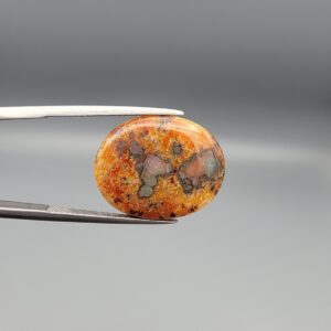 A 10.79-carat oval natural Yemeni carnelian agate cabochon held in tweezers against a gray backdrop. The front side shows a rich, speckled field of red, orange, yellow, and black dendritic inclusions, while the reverse side reveals clear orbicular white-ringed eyes on a translucent golden-orange surface.