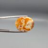 A 10.79-carat oval natural Yemeni carnelian agate cabochon held in tweezers against a gray backdrop. The front side shows a rich, speckled field of red, orange, yellow, and black dendritic inclusions, while the reverse side reveals clear orbicular white-ringed eyes on a translucent golden-orange surface.