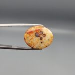 A 12.20-carat oval natural Yemeni carnelian agate cabochon held in tweezers against a gray backdrop. The front displays fine lace-like yellow and orange dendritic patterning with central dark spots, while the reverse side reveals vivid carnelian red, orange, and white cloud formations.