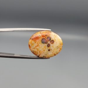 A 12.20-carat oval natural Yemeni carnelian agate cabochon held in tweezers against a gray backdrop. The front displays fine lace-like yellow and orange dendritic patterning with central dark spots, while the reverse side reveals vivid carnelian red, orange, and white cloud formations.