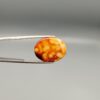 A 15.12-carat oval natural Yemeni carnelian agate cabochon held in tweezers against a neutral gray backdrop. One face shows a bright golden-yellow matrix speckled with dark red and black dendritic inclusions and fine lace edge banding, while the reverse face shows glowing orange and red botryoidal cloud patterns.