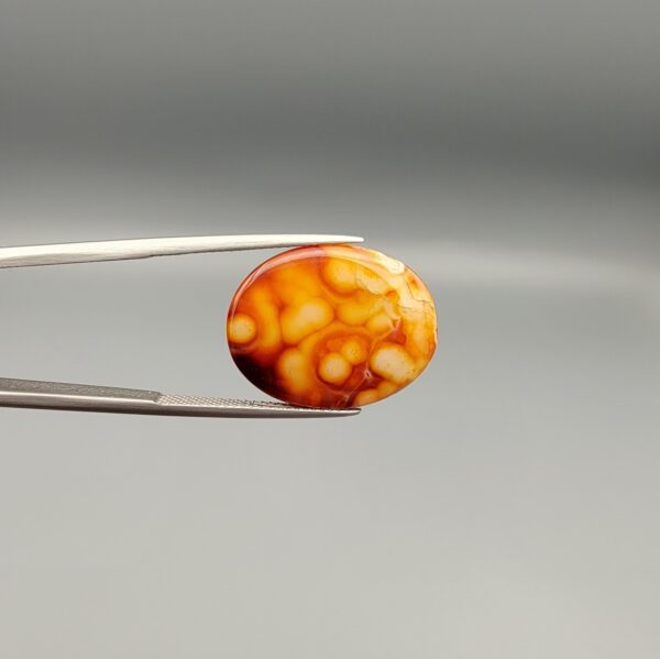 A 15.12-carat oval natural Yemeni carnelian agate cabochon held in tweezers against a neutral gray backdrop. One face shows a bright golden-yellow matrix speckled with dark red and black dendritic inclusions and fine lace edge banding, while the reverse face shows glowing orange and red botryoidal cloud patterns.
