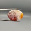 ​An 18.40-carat oval natural Yemeni carnelian agate cabochon held in tweezers against a gray studio background. One face displays a crystalline white-and-red frost matrix edged in bright yellow-orange, while the reverse side reveals a glowing field of carnelian red and orange botryoidal cloud patterns.