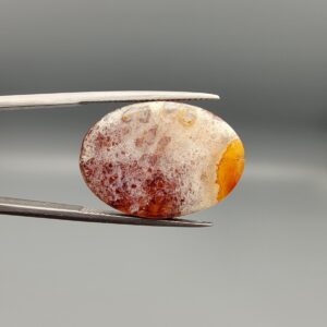 ​An 18.40-carat oval natural Yemeni carnelian agate cabochon held in tweezers against a gray studio background. One face displays a crystalline white-and-red frost matrix edged in bright yellow-orange, while the reverse side reveals a glowing field of carnelian red and orange botryoidal cloud patterns.
