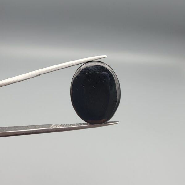 An 18.16-carat oval natural Yemeni agate cabochon held in tweezers against a gray backdrop. The front side shows an ice-white crystalline plume pattern framed by a dark border, while the reverse face is completely solid jet black.