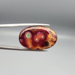 ​A 21.69-carat oval natural Yemeni carnelian agate cabochon held in tweezers against a gray backdrop. The front displays a deep cherry-red surface with orbicular pinkish-white eye rings surrounding a natural green-hued mineral hole, while the reverse side reveals translucent dark carnelian red with golden mossy dendritic plumes.