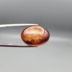 A 14.3-carat oval natural Iranian fire agate cabochon held in metal tweezers against a neutral gray background. One view shows a glowing deep red carnelian base with an exposed golden-white crystalline quartz druzy patch on the surface, while the secondary view displays a smooth red translucent finish.