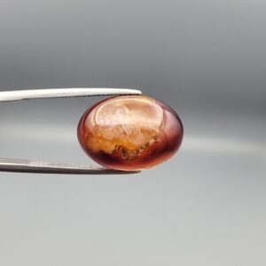 A 14.3-carat oval natural Iranian fire agate cabochon held in metal tweezers against a neutral gray background. One view shows a glowing deep red carnelian base with an exposed golden-white crystalline quartz druzy patch on the surface, while the secondary view displays a smooth red translucent finish.