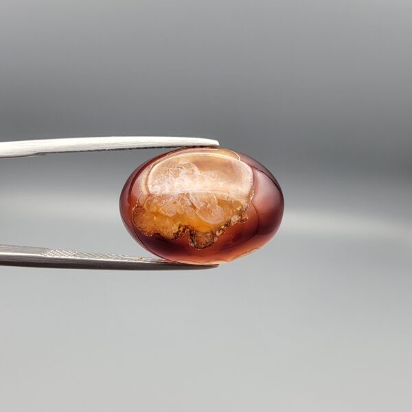A 14.3-carat oval natural Iranian fire agate cabochon held in metal tweezers against a neutral gray background. One view shows a glowing deep red carnelian base with an exposed golden-white crystalline quartz druzy patch on the surface, while the secondary view displays a smooth red translucent finish.
