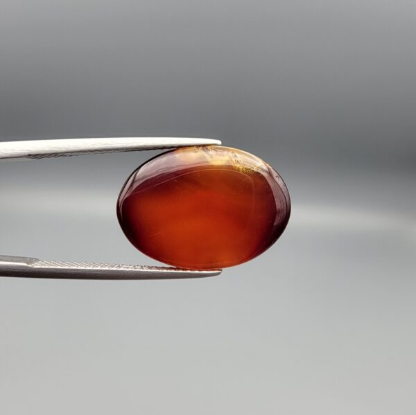A 14.3-carat oval natural Iranian fire agate cabochon held in metal tweezers against a neutral gray background. One view shows a glowing deep red carnelian base with an exposed golden-white crystalline quartz druzy patch on the surface, while the secondary view displays a smooth red translucent finish.