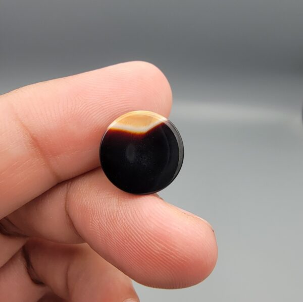 A 12.40-carat precision oval natural Iranian banded agate cabochon resting on a textured black display card. The gemstone features a centered orbicular eye pattern with crisp layers of deep black, white, carnelian red, and chatoyant honey-yellow banding.