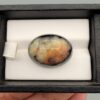 ​A 33.48-carat oval natural Iranian chalcedony agate cabochon displayed in a black presentation box. The high dome features translucent milky agate with multi-colored dendritic inclusions, mossy specks, and geode crystal pocket formations, backed by a seamless, shining jet-black base.
