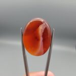 This is a genuine Natural Chalcedony Banded Agate (Sulemani Aqeeq/Hakik) sourced from Iran, weighing 31.80 carats and hand-cut into a smooth oval cabochon. The stone glows a deep, translucent orange-red throughout, with a soft white banded streak curving diagonally through the body and a scattering of fine natural mineral flecks visible beneath the surface. Held to the light, the color deepens and shifts, giving the cabochon real depth rather than a flat, uniform tone. This warm orange-red coloring, distinct from the classic black-and-white Sulemani look, showcases the same natural agate banding process in a fiery, glass-like palette. As part of the broader Sulemani Aqeeq tradition, it carries the same long-standing cultural associations of protection, grounding, and inner strength found across the Middle East and South Asia. The cabochon has been expertly hand-cut and polished to a smooth, glossy finish, ready to be set into a ring, pendant, or custom jewelry piece by your goldsmith or silversmith. As with all natural agate, the banding and color depth are completely unique to this individual stone.
