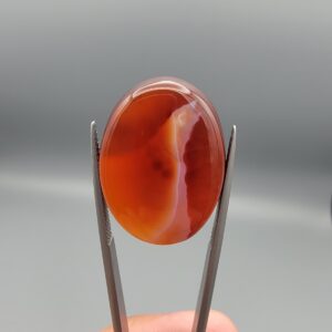 This is a genuine Natural Chalcedony Banded Agate (Sulemani Aqeeq/Hakik) sourced from Iran, weighing 31.80 carats and hand-cut into a smooth oval cabochon. The stone glows a deep, translucent orange-red throughout, with a soft white banded streak curving diagonally through the body and a scattering of fine natural mineral flecks visible beneath the surface. Held to the light, the color deepens and shifts, giving the cabochon real depth rather than a flat, uniform tone. This warm orange-red coloring, distinct from the classic black-and-white Sulemani look, showcases the same natural agate banding process in a fiery, glass-like palette. As part of the broader Sulemani Aqeeq tradition, it carries the same long-standing cultural associations of protection, grounding, and inner strength found across the Middle East and South Asia. The cabochon has been expertly hand-cut and polished to a smooth, glossy finish, ready to be set into a ring, pendant, or custom jewelry piece by your goldsmith or silversmith. As with all natural agate, the banding and color depth are completely unique to this individual stone.