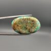 A 41.41-carat oval natural Iranian Neyshabur turquoise cabochon held in metal tweezers against a neutral gray backdrop. The polished front reveals a brilliant greenish-blue tone with intricate golden-brown spiderweb matrix lines, while the unpolished back showcases the raw, natural earth host rock layer.