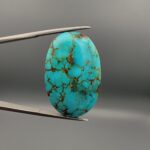 A 41.41-carat oval natural Iranian Neyshabur turquoise cabochon held in metal tweezers against a neutral gray backdrop. The polished front reveals a brilliant greenish-blue tone with intricate golden-brown spiderweb matrix lines, while the unpolished back showcases the raw, natural earth host rock layer.