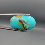 ​A 51.81-carat oval natural Iranian Neyshabur turquoise cabochon held in metal tweezers against a neutral gray background. The smooth top dome shows a rich greenish-blue saturation with dramatic brown and golden matrix veining, while the reverse side reveals its natural, unpolished host rock base.