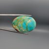 ​A 43.91-carat oval natural Iranian Neyshabur turquoise cabochon held in metal tweezers against a neutral gray backdrop. The polished front shows a vibrant greenish-blue body with swirling golden-brown matrix veining, while the reverse side showcases its unpolished, natural host rock base.