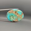 ​A 56.43-carat oval natural Iranian Neyshabur turquoise cabochon held in metal tweezers against a neutral gray background. The smooth, glossy front face displays a rich, vibrant greenish-blue color with fine hairline matrix veins, while the reverse side reveals raw, unpolished host rock integrated with turquoise pockets.