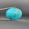 ​A 56.43-carat oval natural Iranian Neyshabur turquoise cabochon held in metal tweezers against a neutral gray background. The smooth, glossy front face displays a rich, vibrant greenish-blue color with fine hairline matrix veins, while the reverse side reveals raw, unpolished host rock integrated with turquoise pockets.