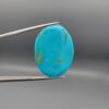 ​An 18.12-carat vertical oval natural Iranian Neyshabur turquoise cabochon held in metal tweezers against a neutral gray background. The front presents a smooth, vibrant greenish-blue dome with fine golden-brown matrix accents, while the reverse side reveals its natural, unpolished earth matrix base.