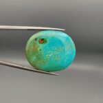 A 31.99-carat oval natural Iranian Neyshabur turquoise cabochon held in metal tweezers against a neutral gray backdrop. The polished front dome features a rich greenish-blue tone with fine golden-brown matrix mottling and webbed textures, while the back reveals a raw, unpolished earth host-rock base.