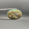 A 24.92-carat oval natural Iranian Neyshabur turquoise cabochon held in metal tweezers against a neutral gray background. The front presents a smooth greenish-blue dome with intricate golden-brown spiderweb veining, while the reverse side showcases its unpolished, natural host-rock base.