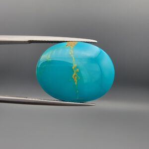 ​A 21.19-carat oval natural Iranian Neyshabur turquoise cabochon held in metal tweezers against a neutral gray background. The front shows a smooth, high-lustre greenish-blue dome with a single vertical golden-brown matrix line running across the center, while the reverse side reveals a natural, unpolished host-rock backing.