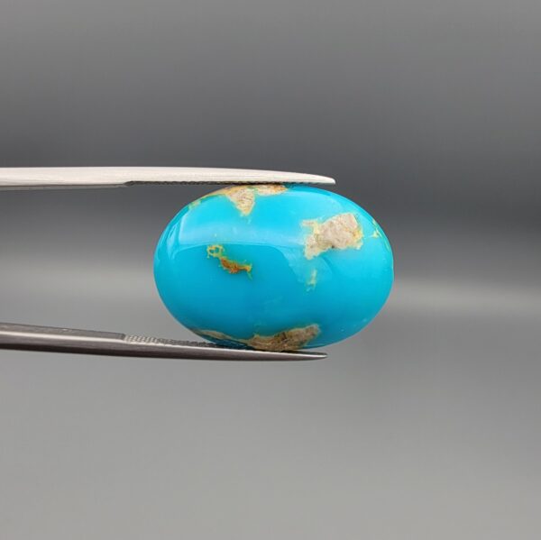A 22.36-carat oval natural Iranian Neyshabur turquoise cabochon held in metal tweezers against a neutral gray background. The front exhibits a rich sky-blue tone with soft brownish-white natural matrix patches, while the reverse side showcases a raw brown matrix core interwoven with blue turquoise.
