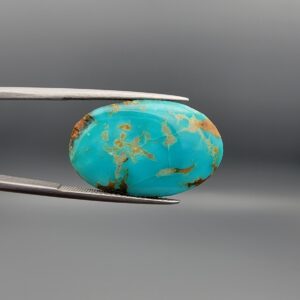 A 21.23-carat oval natural Iranian Neyshabur turquoise cabochon held in metal tweezers against a neutral gray background. The face shows a bright greenish-blue dome with natural golden-brown matrix textures and an authentic mountain fissure mark on the left edge, while the reverse side reveals a raw brown host-rock base.