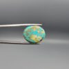 ​A 17.29-carat oval natural Iranian Neyshabur turquoise cabochon held in metal tweezers against a neutral gray background. The front presents a smooth greenish-blue dome with naturally distributed golden-brown matrix mottling, while the reverse side reveals a flat surface covered with golden host rock and turquoise patterning.