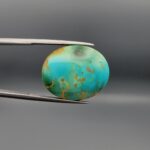 A 22.46-carat oval natural Iranian Neyshabur turquoise cabochon held in metal tweezers against a neutral gray background. The front showcases a smooth greenish-blue dome with a naturally formed network of golden-brown matrix lines, while the reverse side reveals a flat, unpolished light host-rock base.