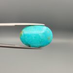 ​A 15.84-carat oval natural Iranian Neyshabur turquoise cabochon held in metal tweezers against a neutral gray background. The front presents a smooth greenish-blue dome with faint brownish matrix highlights, while the reverse side reveals raw earthy host rock featuring a natural mountain cavity formed during extraction.