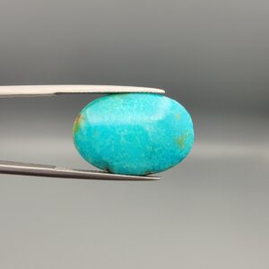 ​A 15.84-carat oval natural Iranian Neyshabur turquoise cabochon held in metal tweezers against a neutral gray background. The front presents a smooth greenish-blue dome with faint brownish matrix highlights, while the reverse side reveals raw earthy host rock featuring a natural mountain cavity formed during extraction.