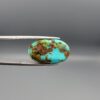 A 14.47-carat oval natural Iranian Neyshabur turquoise cabochon held in metal tweezers against a neutral gray background. The front presents a high-gloss greenish-blue dome with sparse, naturally formed brown matrix flecks, while the reverse side reveals a flat surface showing earthy brown host rock intermixed with raw turquoise.