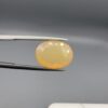 Studio photograph held in steel tweezers showcasing a 5.79 carat natural oval-shaped Ethiopian opal loose gemstone with a double-side fancy cut featuring flat and domed facets.