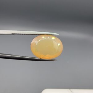 Studio photograph held in steel tweezers showcasing a 5.79 carat natural oval-shaped Ethiopian opal loose gemstone with a double-side fancy cut featuring flat and domed facets.