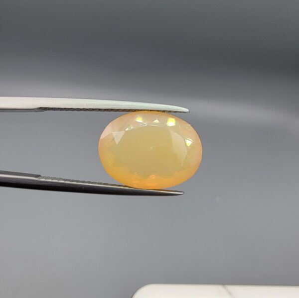 Studio photograph held in steel tweezers showcasing a 5.79 carat natural oval-shaped Ethiopian opal loose gemstone with a double-side fancy cut featuring flat and domed facets.