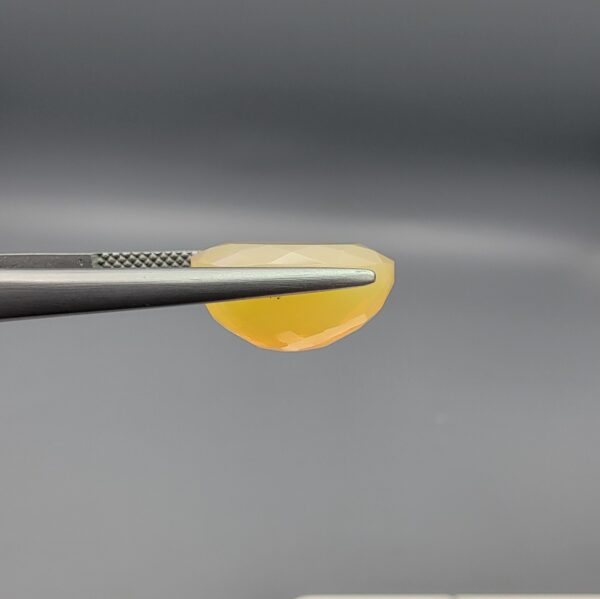 Studio photograph held in steel tweezers showcasing a 5.79 carat natural oval-shaped Ethiopian opal loose gemstone with a double-side fancy cut featuring flat and domed facets.